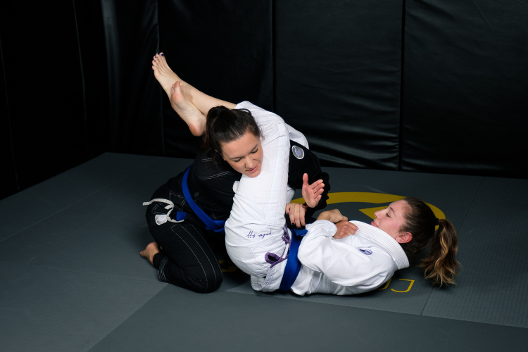 Two women practicing Brazilian Jiu-Jitsu on a padded mat, one applying a submission hold while both wear training gis and blue belts.