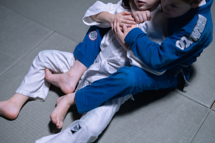 Two children practicing Brazilian Jiu-Jitsu on a mat, one applying a rear choke position while both wear training gis.