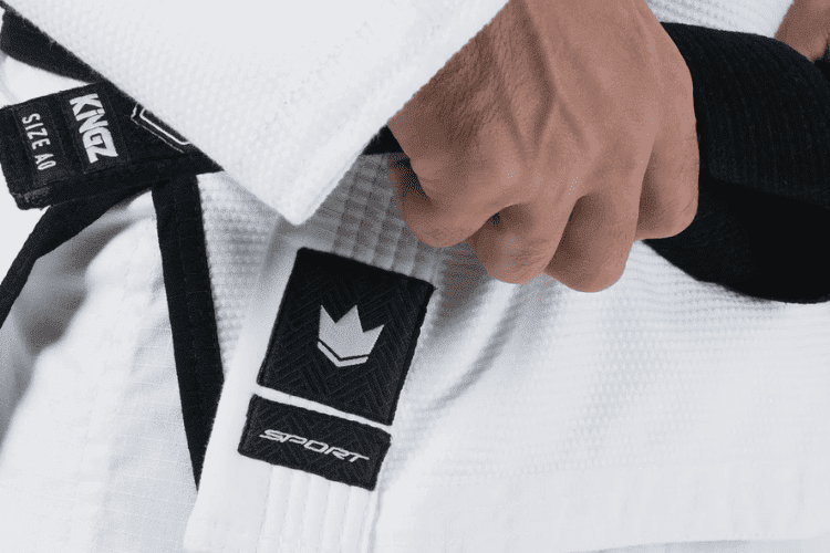 Close-up of a martial artist in a white gi tying a black belt with visible Kingz Kimonos branding.